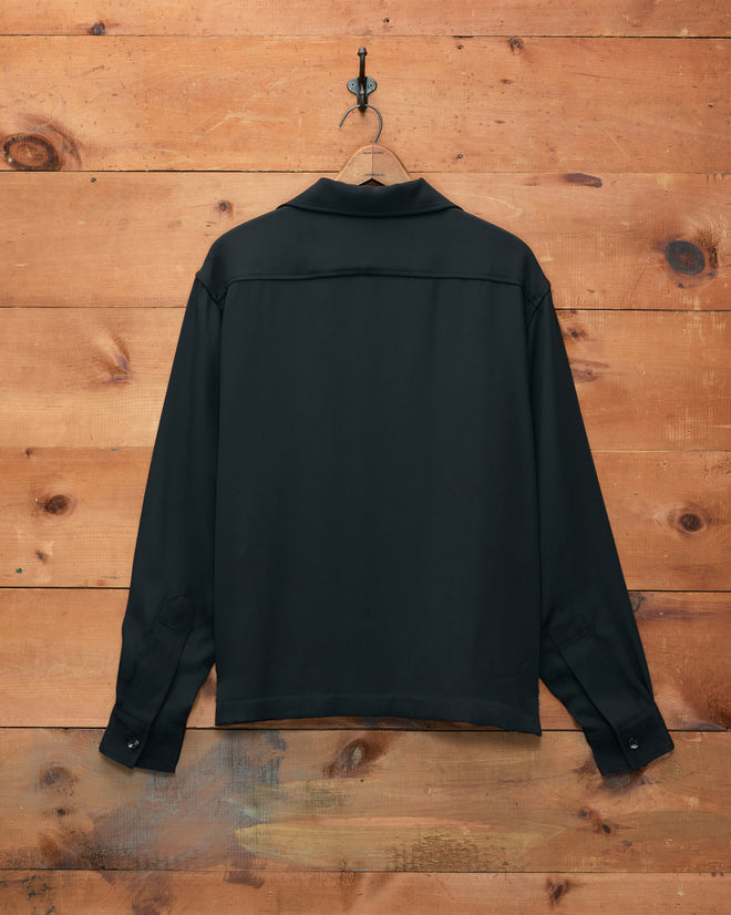 The Trillium Long Sleeve Shirt - Black, crafted from silk crepe in a 1940s-inspired men’s silhouette, hangs on a hook against a wooden wall, displayed from the back.