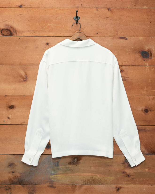 The Trillium Long Sleeve Shirt in cream, crafted from white silk crepe, hangs on a hook against a wooden wall and is shown from the back. It features subtle trompe l’oeil frog closures inspired by 1940s men’s shirts.
