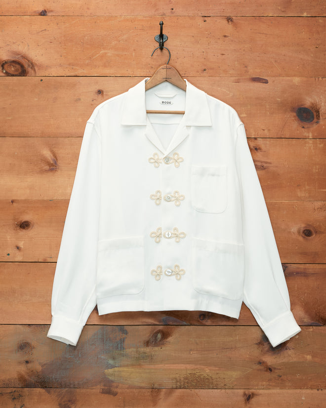 Trillium Long Sleeve Shirt - Cream, featuring decorative trompe l’oeil frog closures and three pockets inspired by 1940s men’s shirts, hangs on a wooden wall.