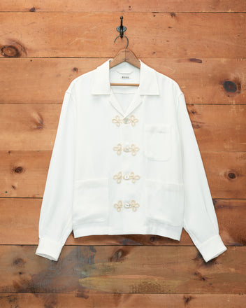 Trillium Long Sleeve Shirt - Cream, featuring decorative trompe l’oeil frog closures and three pockets inspired by 1940s men’s shirts, hangs on a wooden wall.