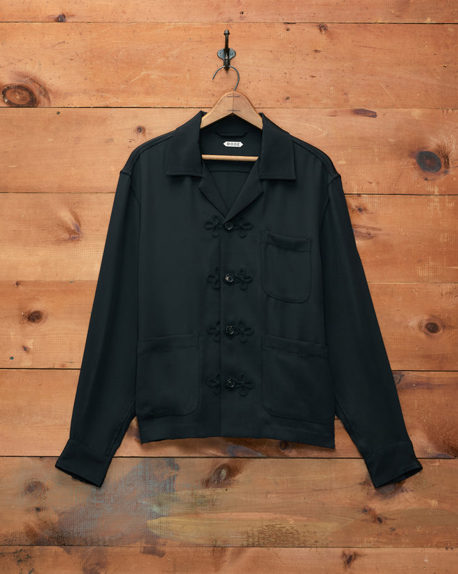 Trillium Long Sleeve Shirt - Black features a button-up design with three front pockets, hanging on a hook against a wooden wall background. Reminiscent of 1940s men’s shirts, it is finished with subtle trompe l’oeil frog closures that add vintage charm.