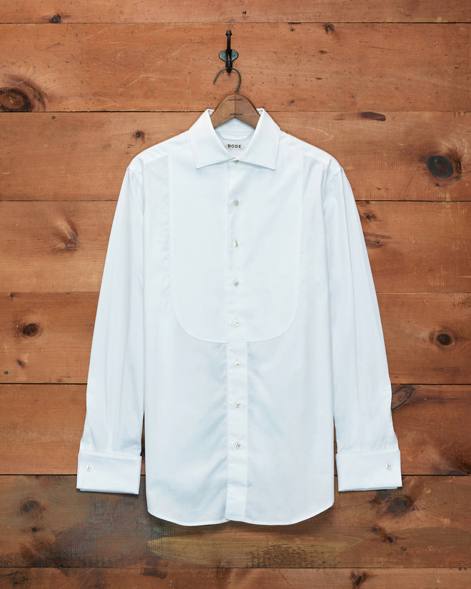 A Bib Tuxedo Shirt in crisp white piqué cotton with button cuffs hangs on a hook against a rustic wooden wall.