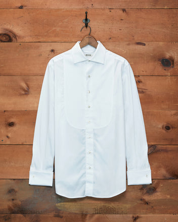 A Bib Tuxedo Shirt in crisp white piqué cotton with button cuffs hangs on a hook against a rustic wooden wall.