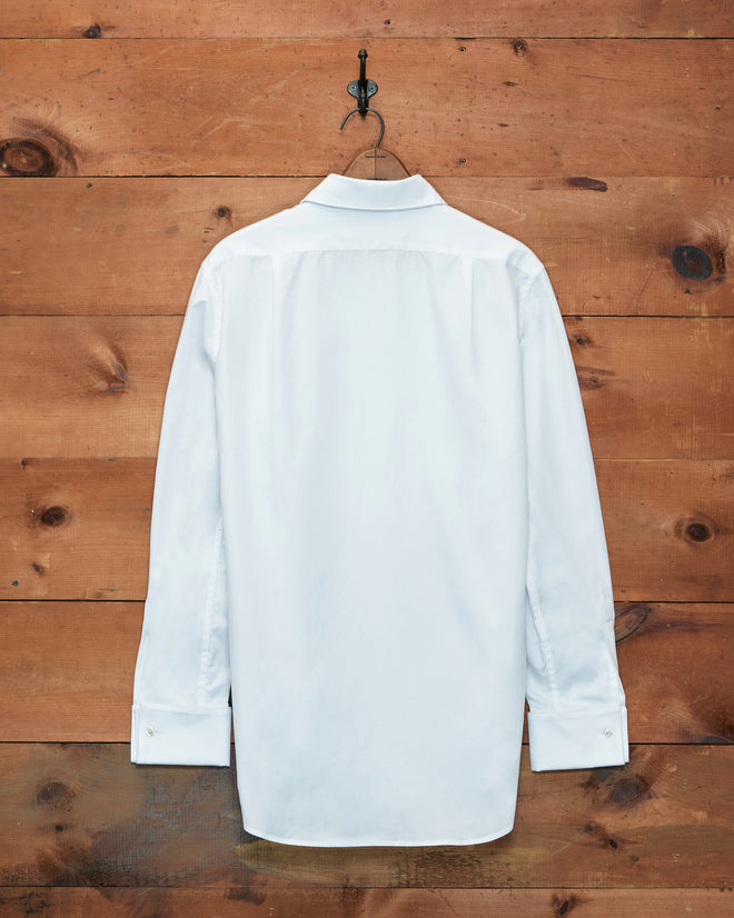 The Bib Tuxedo Shirt, crafted from white piqué cotton, hangs on a hook against a wooden wall and is shown from the back.