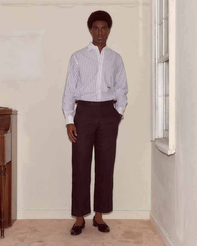 A person stands indoors near a window, wearing a striped shirt and Lightweight Linen Side-Buckle Trousers - Black with black shoes.