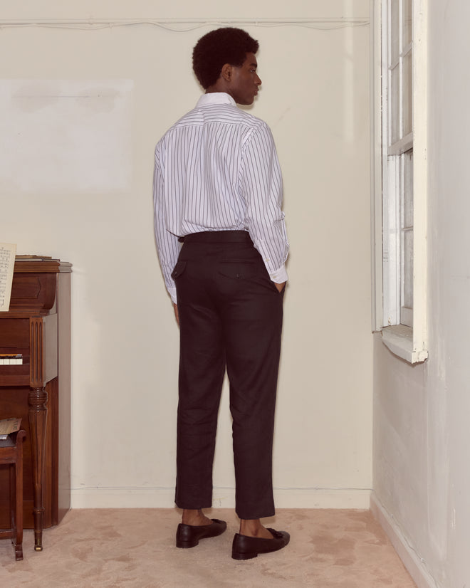 A person in a striped shirt and Lightweight Linen Side-Buckle Trousers - Black stands facing a wall near a piano and a window.
