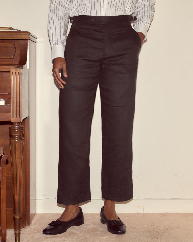 A person wearing Lightweight Linen Side-Buckle Trousers - Black, a striped shirt, and black loafers stands indoors next to a wooden piano.