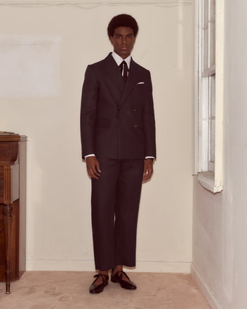 A man wearing the Lightweight Linen Double-Breasted Suit Jacket in black with cropped pants stands indoors by a window, looking at the camera.