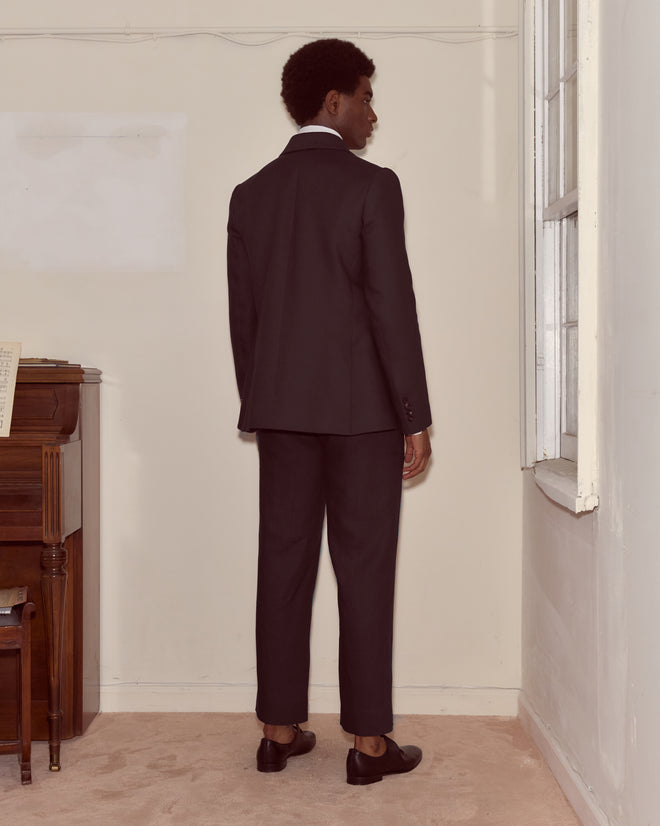 A man wearing the Lightweight Linen Double-Breasted Suit Jacket in black stands facing away in a room with a piano and a large window.