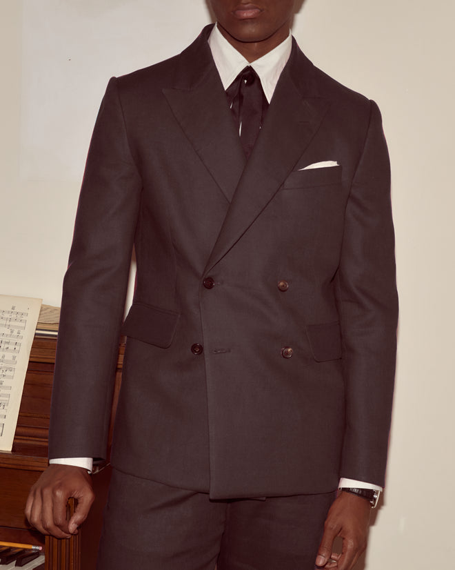 A person in a Lightweight Linen Double-Breasted Suit Jacket - Black, paired with a white shirt and black tie, stands by a piano with sheet music.