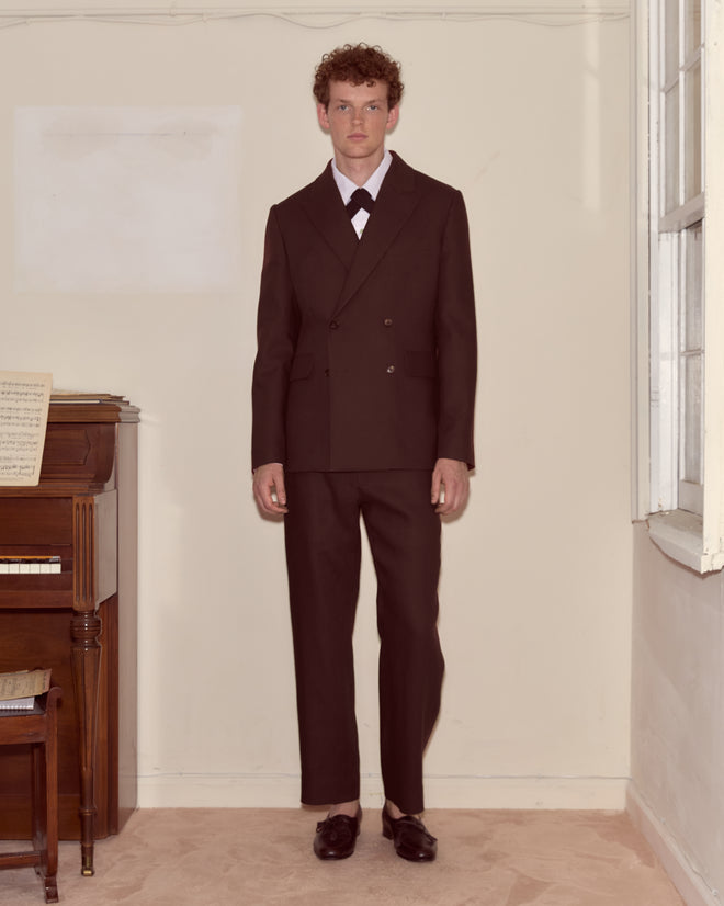 A man wearing the Lightweight Linen Double-Breasted Suit Jacket in Brown/Black stands indoors by a piano and window, facing forward with a neutral expression.