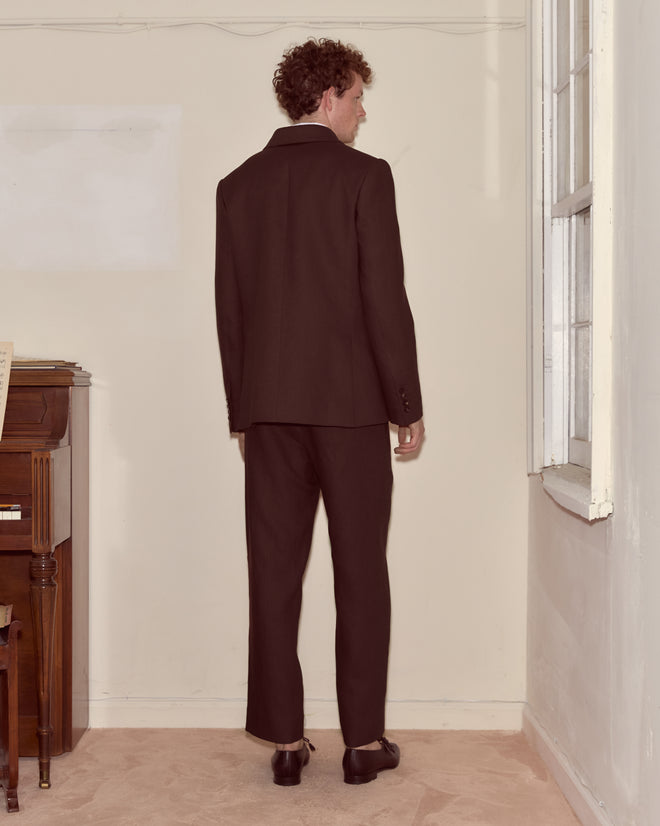 A man wearing the Lightweight Linen Double-Breasted Suit Jacket in Brown/Black stands by a beige wall near a window and a wooden piano.