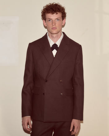 A young man with curly hair wears the Lightweight Linen Double-Breasted Suit Jacket in brown/black, standing against a plain light-colored wall.