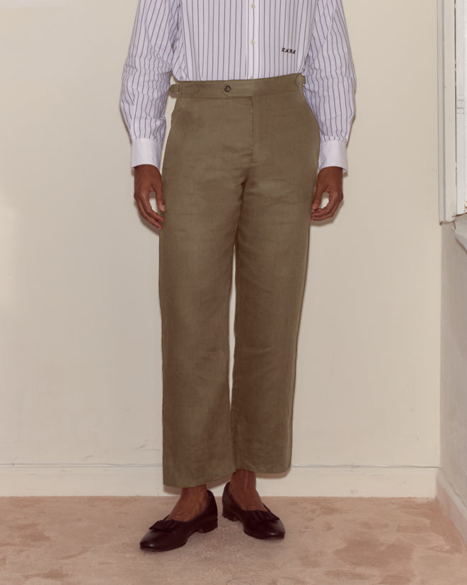 A person wearing Lightweight Linen Side-Buckle Trousers in olive, a striped shirt, and dark loafers stands against a cream-colored wall.