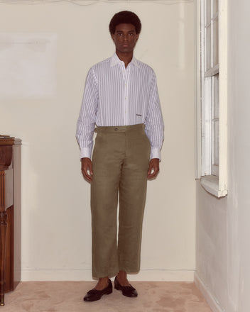 A man in a striped shirt and Lightweight Linen Side-Buckle Trousers - Olive stands indoors by a window, facing the camera.