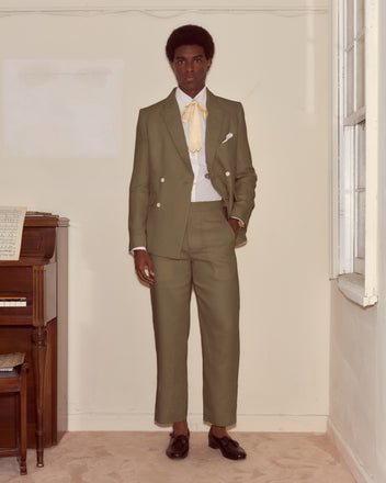 A man in a Lightweight Linen Double-Breasted Suit Jacket - Olive with a yellow tie stands indoors by a piano and window, showcasing effortless spring-summer style.