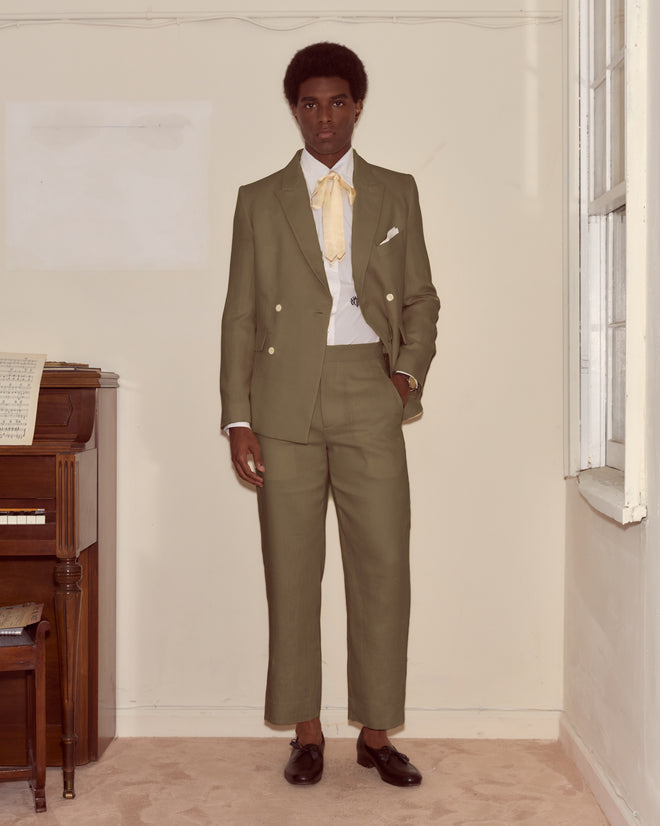 A man in a Lightweight Linen Double-Breasted Suit Jacket - Olive with a yellow tie stands indoors by a piano and window, showcasing effortless spring-summer style.