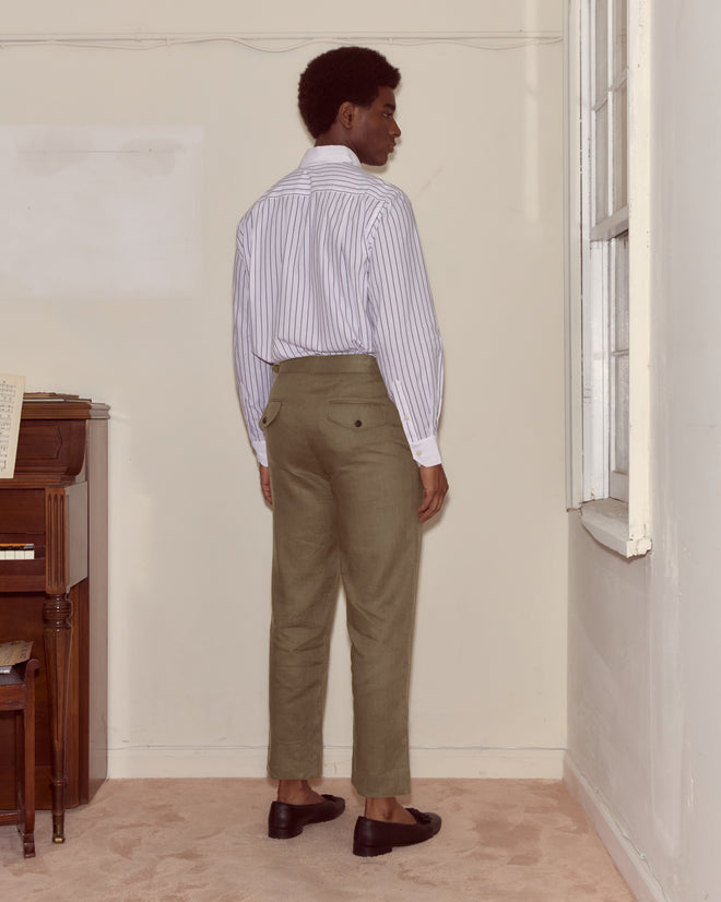 A person stands indoors facing away, wearing a striped shirt, black shoes, and Lightweight Linen Side-Buckle Trousers in olive.