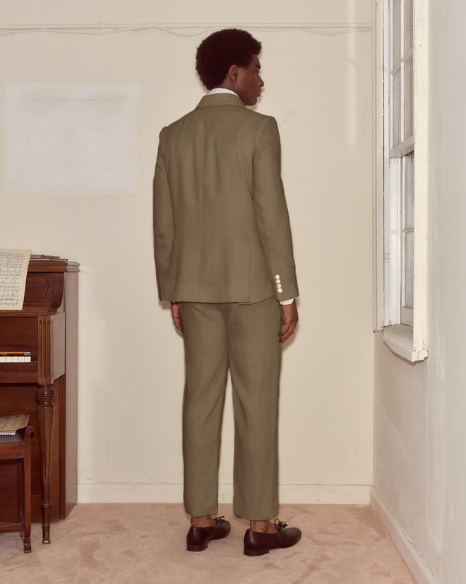 A person in the Lightweight Linen Double-Breasted Suit Jacket - Olive stands facing a wall in a room with a piano and sheet music on the left, ideal for spring and summer style.