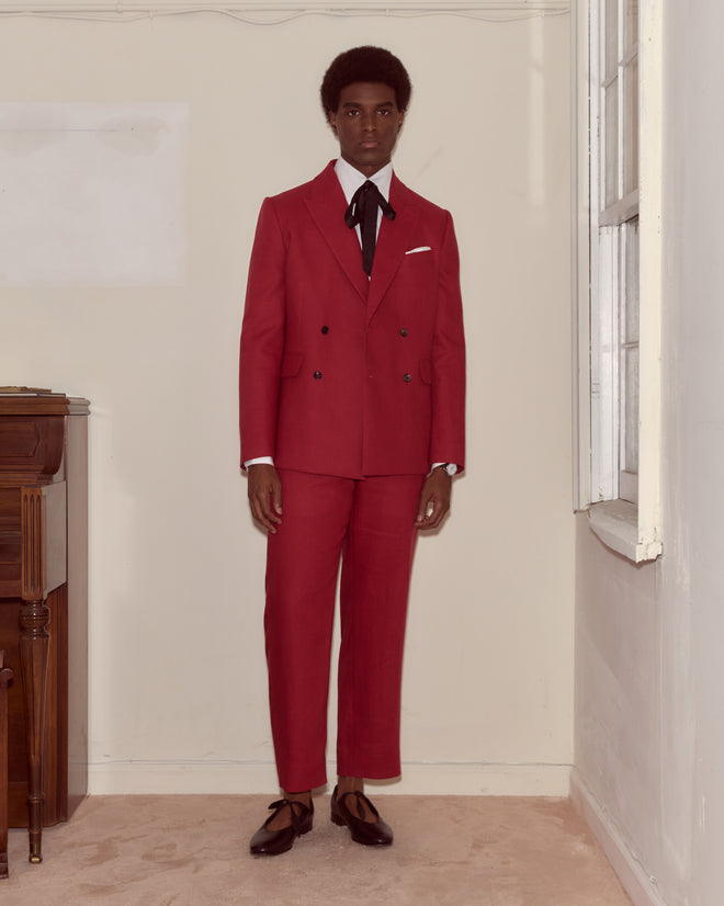A person wearing the Lightweight Linen Double-Breasted Suit Jacket in red with a black tie stands indoors by a window and wooden table, showcasing perfect spring-summer style.