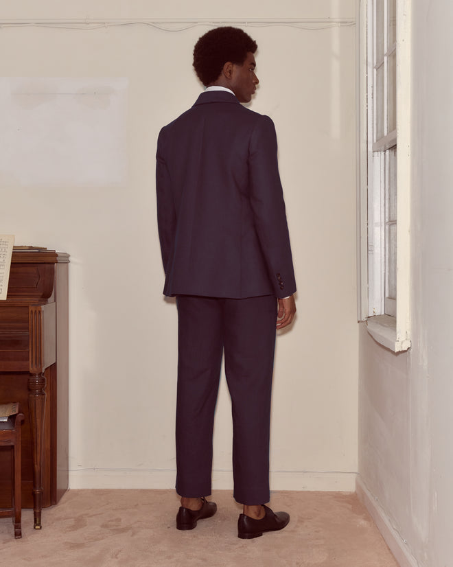 A man in the Lightweight Linen Double-Breasted Suit Jacket - Midnight stands indoors by a piano and window, facing the wall.