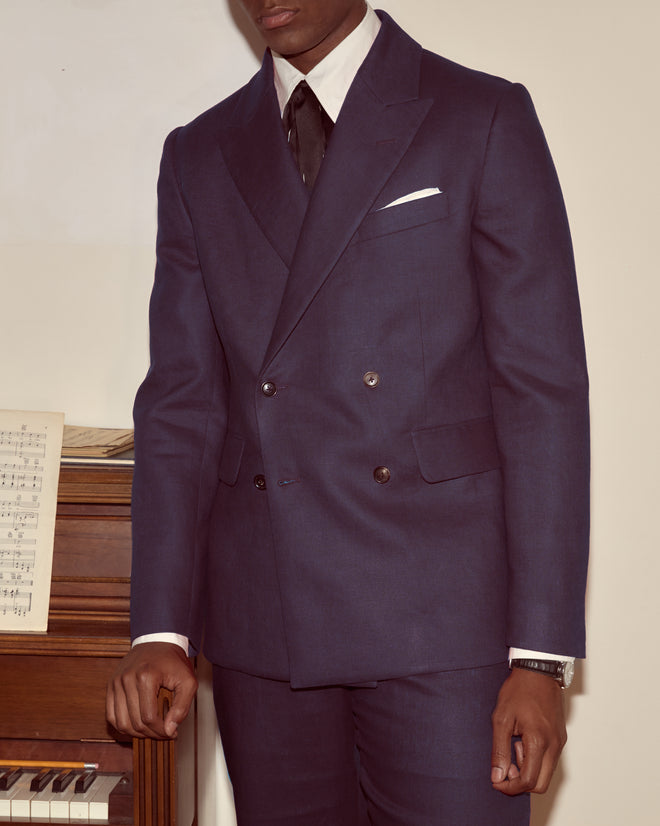 A man in a Lightweight Linen Double-Breasted Suit Jacket - Midnight stands by a piano with sheet music, only his upper body visible.