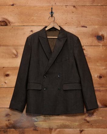 The Lightweight Linen Double-Breasted Suit Jacket in Brown/Black is displayed on a wooden hanger against a rustic wooden wall.