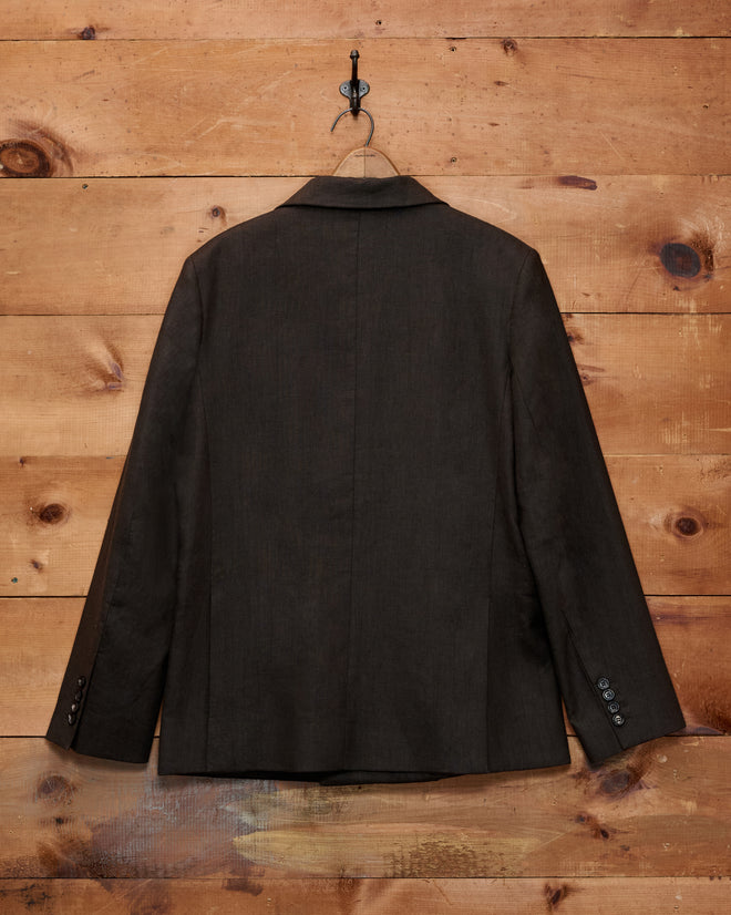 A Lightweight Linen Double-Breasted Suit Jacket in brown/black hangs on a hook against a wooden wall, shown from the back.