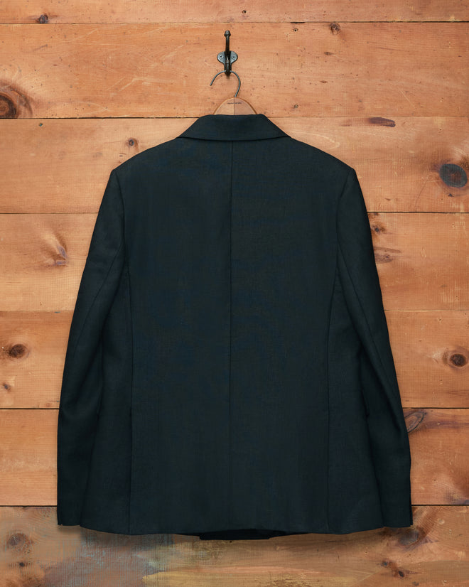 The Lightweight Linen Double-Breasted Suit Jacket in black, displayed from the back, hangs on a hook against a wooden wall.