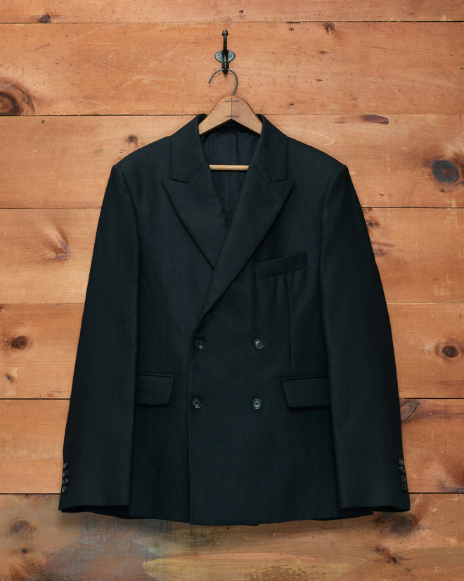 The Lightweight Linen Double-Breasted Suit Jacket in black, ideal as a men’s spring jacket, hangs on a wooden hanger against a wooden wall.