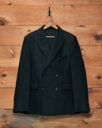 The Lightweight Linen Double-Breasted Suit Jacket in black, ideal as a men’s spring jacket, hangs on a wooden hanger against a wooden wall.