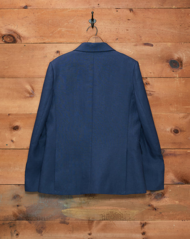 A Lightweight Linen Double-Breasted Suit Jacket in Midnight hangs on a hook against a wooden wall.