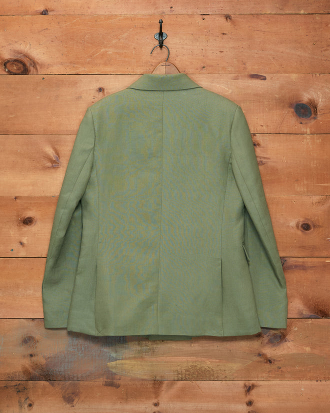 A Lightweight Linen Double-Breasted Suit Jacket in olive hangs on a black hook against a wooden wall background, perfect for men’s spring suiting.