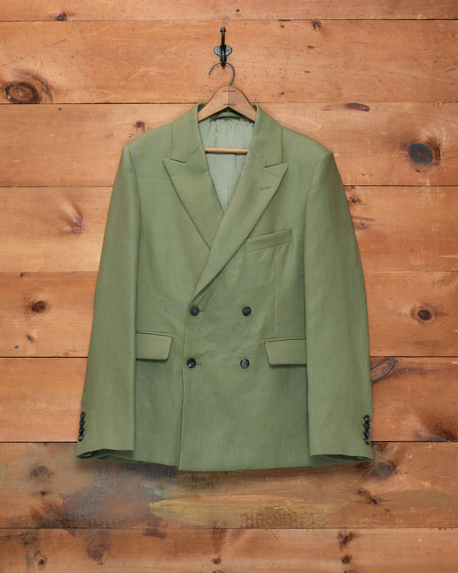 The Lightweight Linen Double-Breasted Suit Jacket in olive, ideal for men’s spring suiting, hangs on a wooden hanger against a wooden wall.