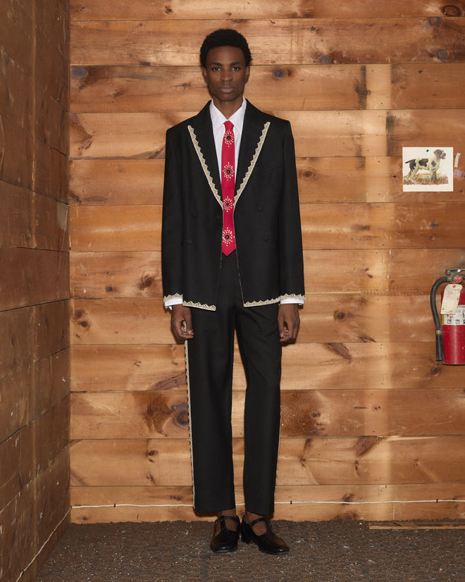 Man in Lacework Side-Buckle Trousers featuring cream machine lace trim and a black suit, with a red tie, stands in front of a wood-paneled wall and a fire extinguisher.
