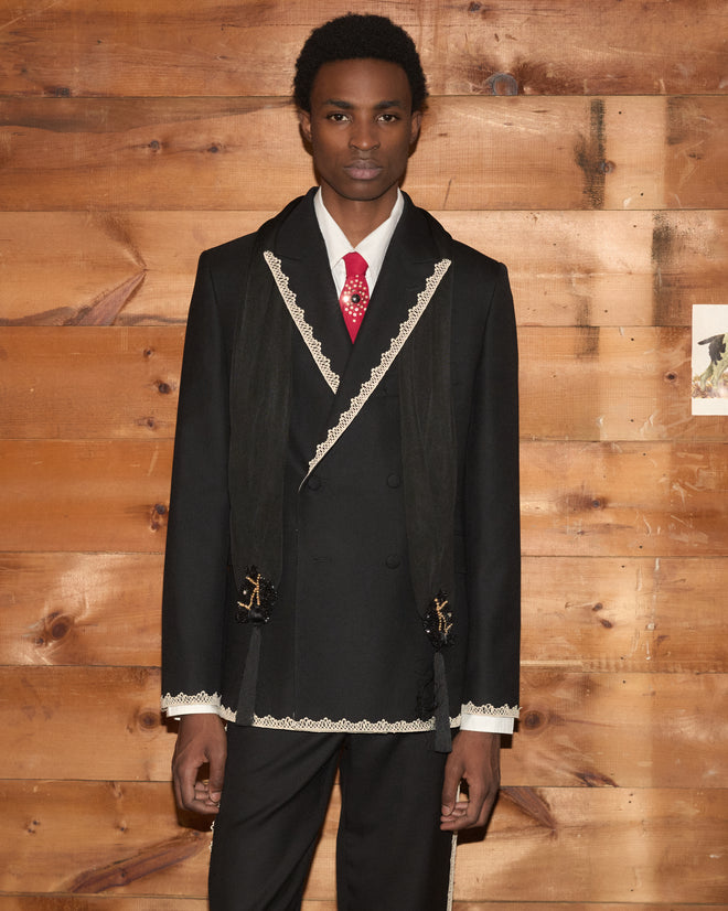 A man in a Lacework Double-Breasted Tuxedo Jacket with white trim and a red tie stands in front of a wooden wall.