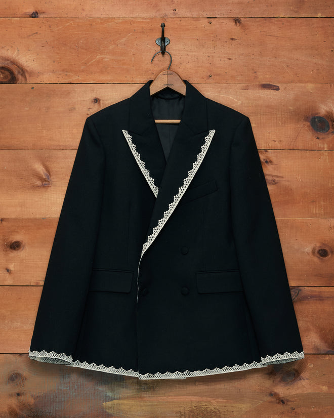 The Lacework Double-Breasted Tuxedo Jacket, featuring black fabric with white machine lace trim on the lapels and hem in a style reminiscent of a classic midcentury tuxedo, is displayed hanging on a wooden wall.