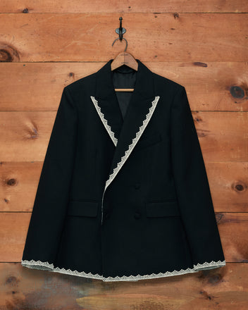 The Lacework Double-Breasted Tuxedo Jacket, featuring black fabric with white machine lace trim on the lapels and hem in a style reminiscent of a classic midcentury tuxedo, is displayed hanging on a wooden wall.