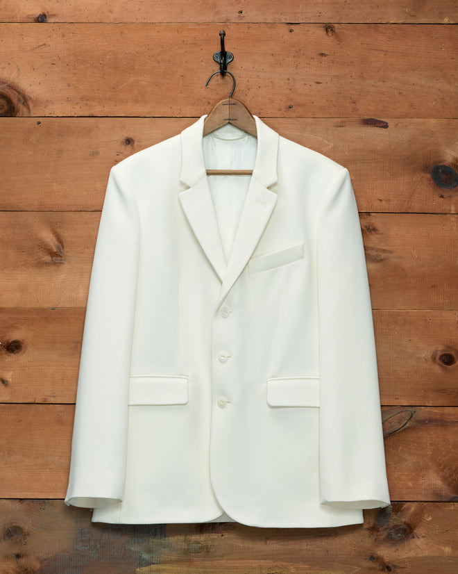 Cream-colored Riding Twill Suit Jacket with three buttons, hanging on a wooden hanger against a wood-paneled wall.