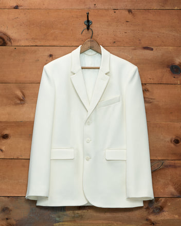 Cream-colored Riding Twill Suit Jacket with three buttons, hanging on a wooden hanger against a wood-paneled wall.