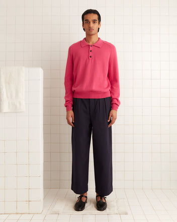 A person stands in a white-tiled room, wearing a long-sleeve pink cashmere polo sweater, dark pleated pants, and black shoes. They face the camera with a neutral expression.