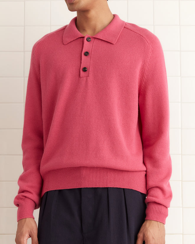 A person stands in a white-tiled room, wearing a long-sleeve pink cashmere polo sweater, dark pleated pants, and black shoes. They face the camera with a neutral expression.