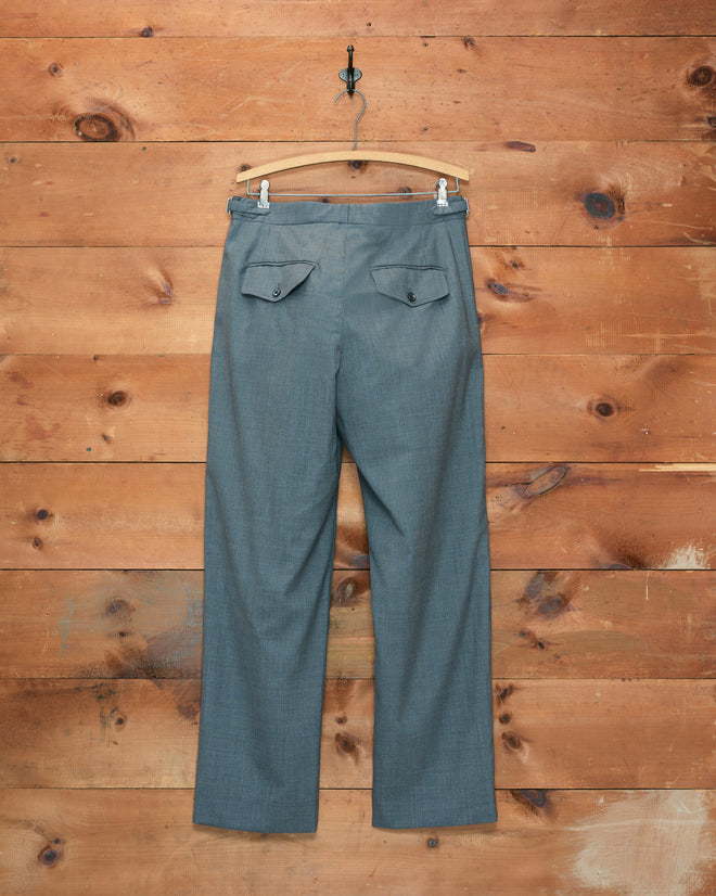 The Tropical Wool Side-Buckle Trousers - Grey are displayed on a wooden hanger, set against a rustic wooden plank wall.
