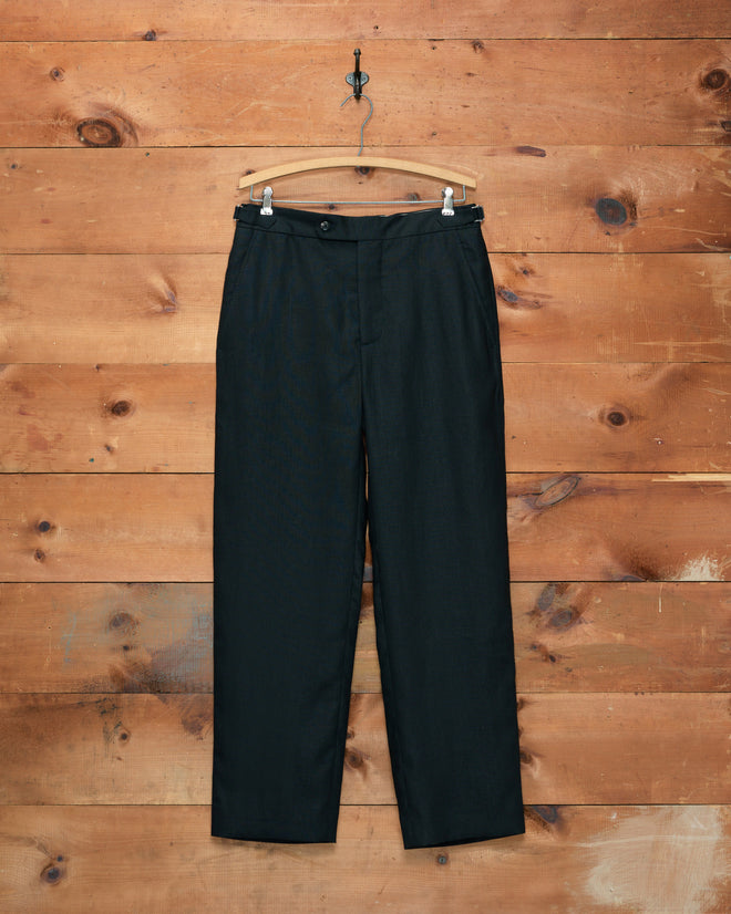 Lightweight Linen Side-Buckle Trousers - Black hang elegantly on a wooden hanger against a rustic wooden wall background, seamlessly blending timeless style with breathable comfort for the warmer months.