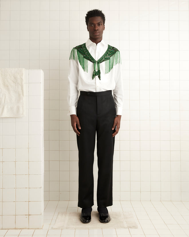 The Geode Collar - Green, with its intricate beadwork and fringe detail, hangs on a wooden hanger against a white tiled wall.