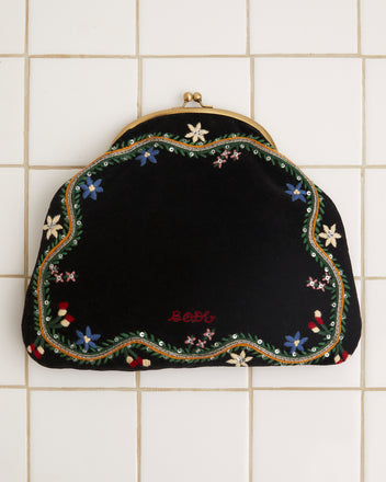 The Alpineflower Clutch, a vintage black velvet piece adorned with an embroidered wildflower pattern and a metal clasp, rests gracefully on a white tiled background.