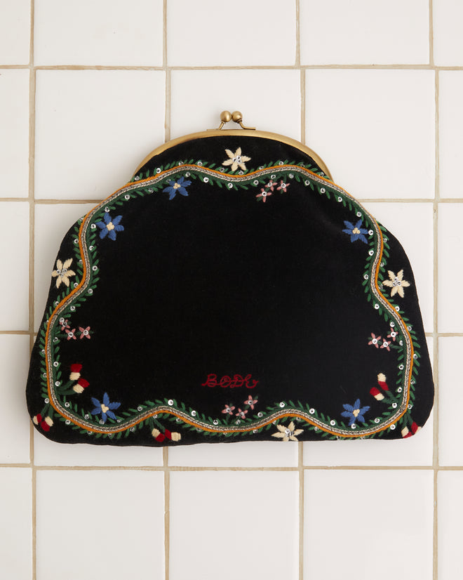 The Alpineflower Clutch, a vintage black velvet piece adorned with an embroidered wildflower pattern and a metal clasp, rests gracefully on a white tiled background.