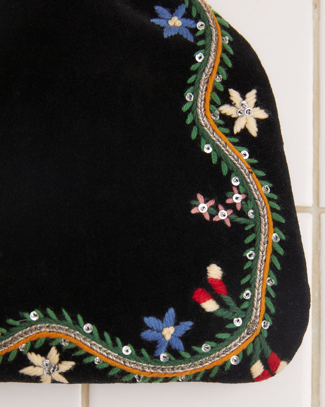 The Alpineflower Clutch, a vintage black velvet piece adorned with an embroidered wildflower pattern and a metal clasp, rests gracefully on a white tiled background.