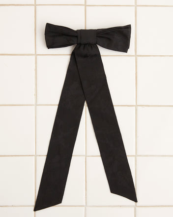 A large Football Jacquard Bow Barrette in black, featuring long tails and crafted from vintage-inspired deadstock woven fabric, is displayed on a white tiled surface.