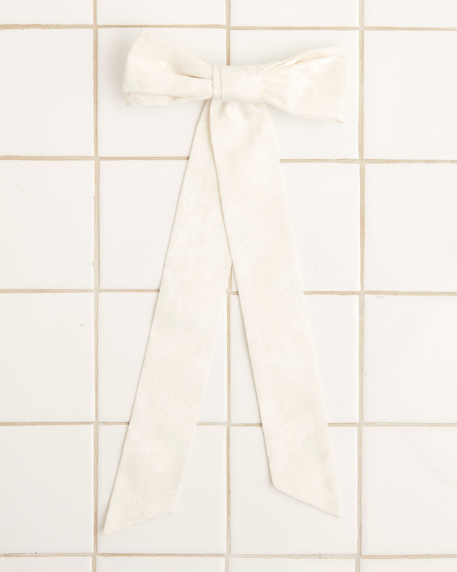 The Football Jacquard Bow Barrette in cream, which channels the vintage style of the 1950s-1960s with its long tails, is displayed on a white tiled background.